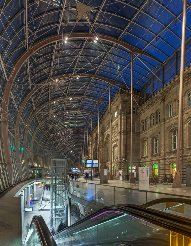 Inside the glass canopy of Strasbourg station, the lit sandstone facade behind the glass